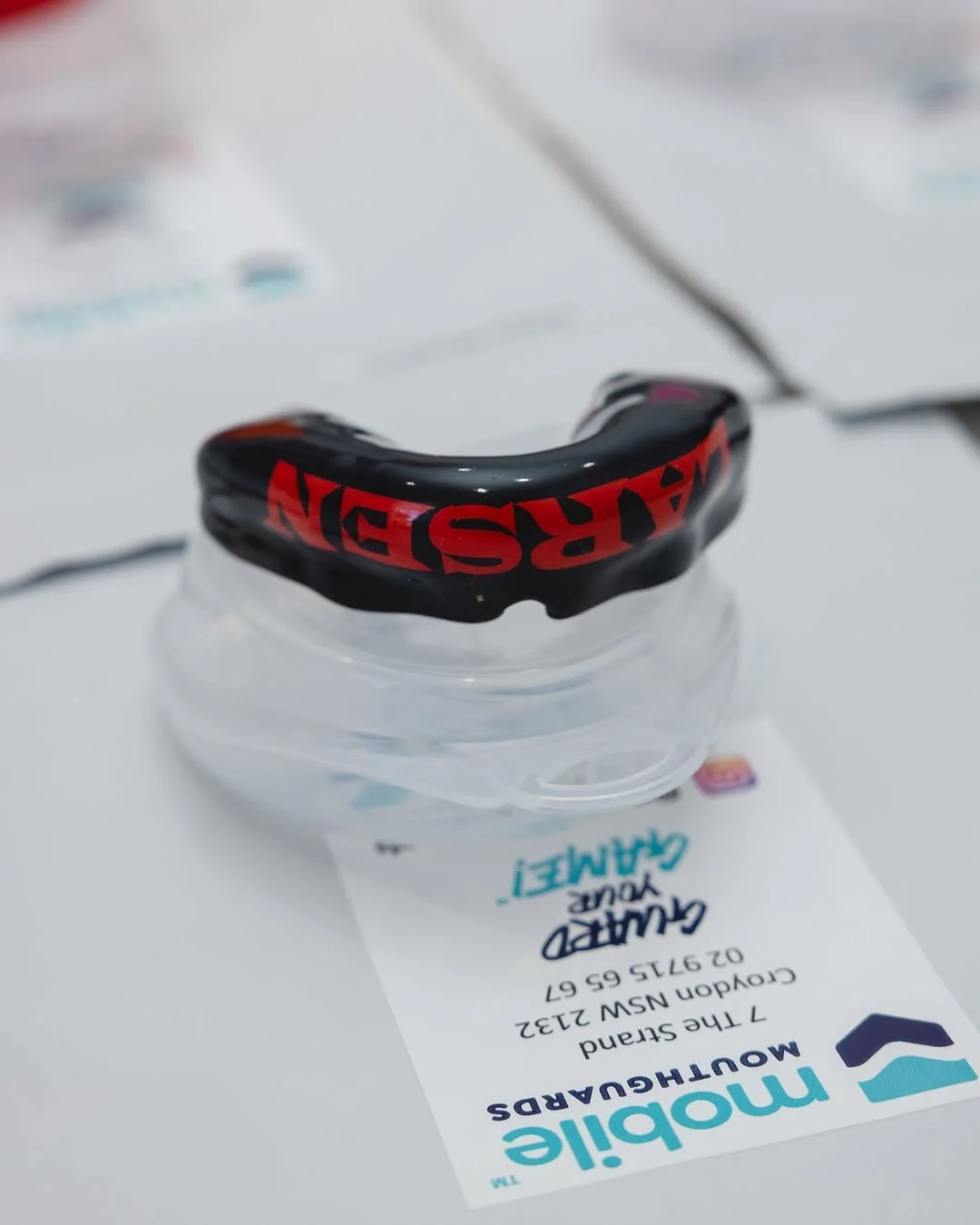 A custom black sports mouthguard with red lettering sits on its clear case next to a clinic card.