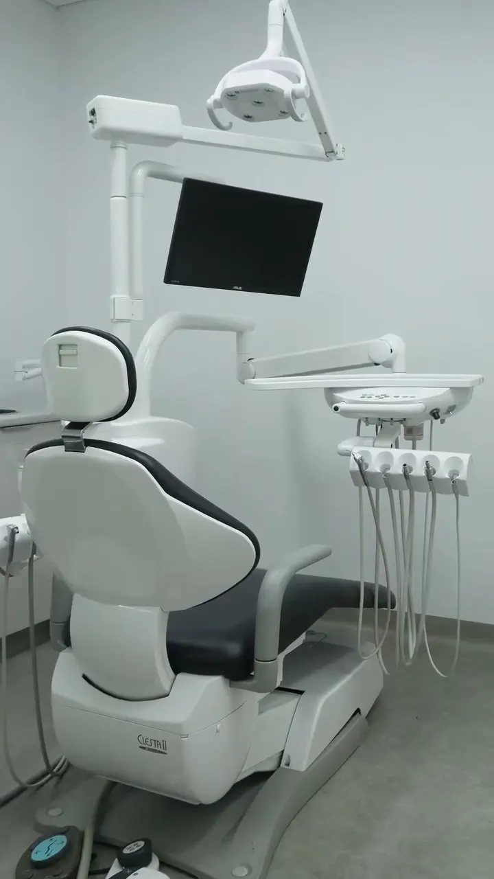 An empty treatment room with a white and grey dental chair, overhead light and monitor
