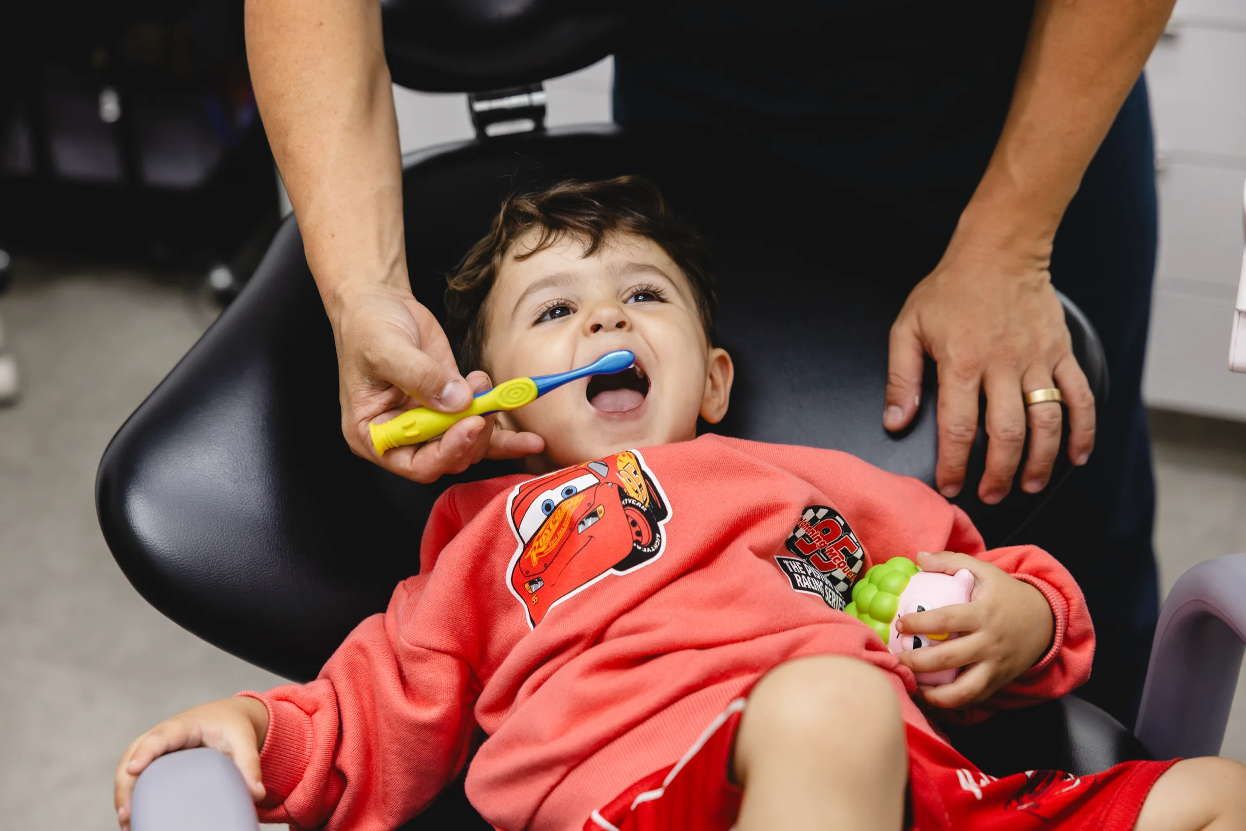 A laughing toddler brushing his teeth in the dental chair, with a grown-up holding the brush